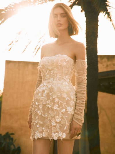 Model draagt de Pronovias Lulu korte trouwjurk met pailletten en glitter tule onder palmbomen.